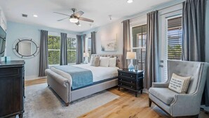A bedroom with a large bed, a ceiling fan, a nightstand, a chair, and a window with curtains.