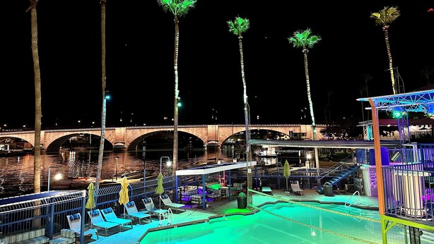 Vista nocturna de un área de piscina con sillones de descanso y sombrillas, un puente al fondo y palmeras.