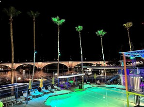A nighttime view of a pool area with lounge chairs and umbrellas, a bridge in the background, and palm trees. - DESERT PARADISE - dreamy pool, lagoon waterfall, hot tub, & stunning lake views (Lake Havasu City)