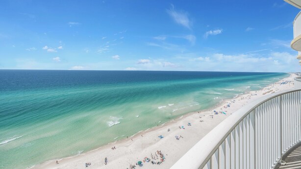 Blick auf einen Strand von einem Balkon mit Geländer, strahlend blauer Himmel und türkisfarbenes Meer.