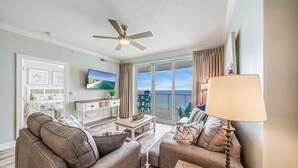 Ein Wohnzimmer mit Deckenventilator, Fernseher, Sofa, Couchtisch und Blick ins Freie durch große Fenster.