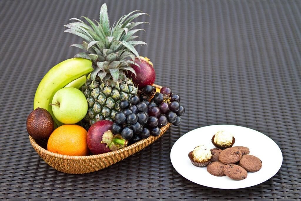 A fruit basket with bananas, apples, grapes, and a pineapple on a woven table.