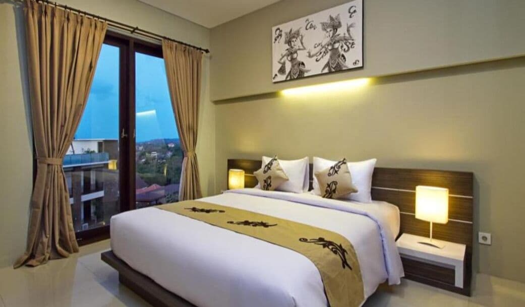 A hotel room with a bed, two bedside tables with lamps, a large window with curtains, and a wall-mounted artwork.