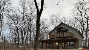 Una baita con balcone e portico coperto, circondata da alberi e terreno ricoperto di foglie.