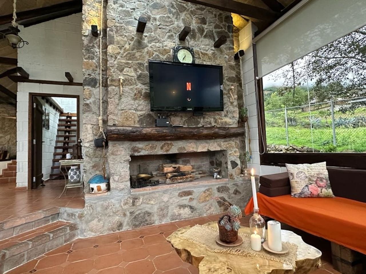 Ein Kamin aus Stein mit darüber montiertem Fernseher, eine Sitzecke mit Kissen und Blick ins Grüne.