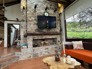 A stone fireplace with a television mounted above it, a seating area with cushions, and a view of greenery outside. - Villa Nathalie, jaccuzzi, king size bed+hammock (Chican, Paute)