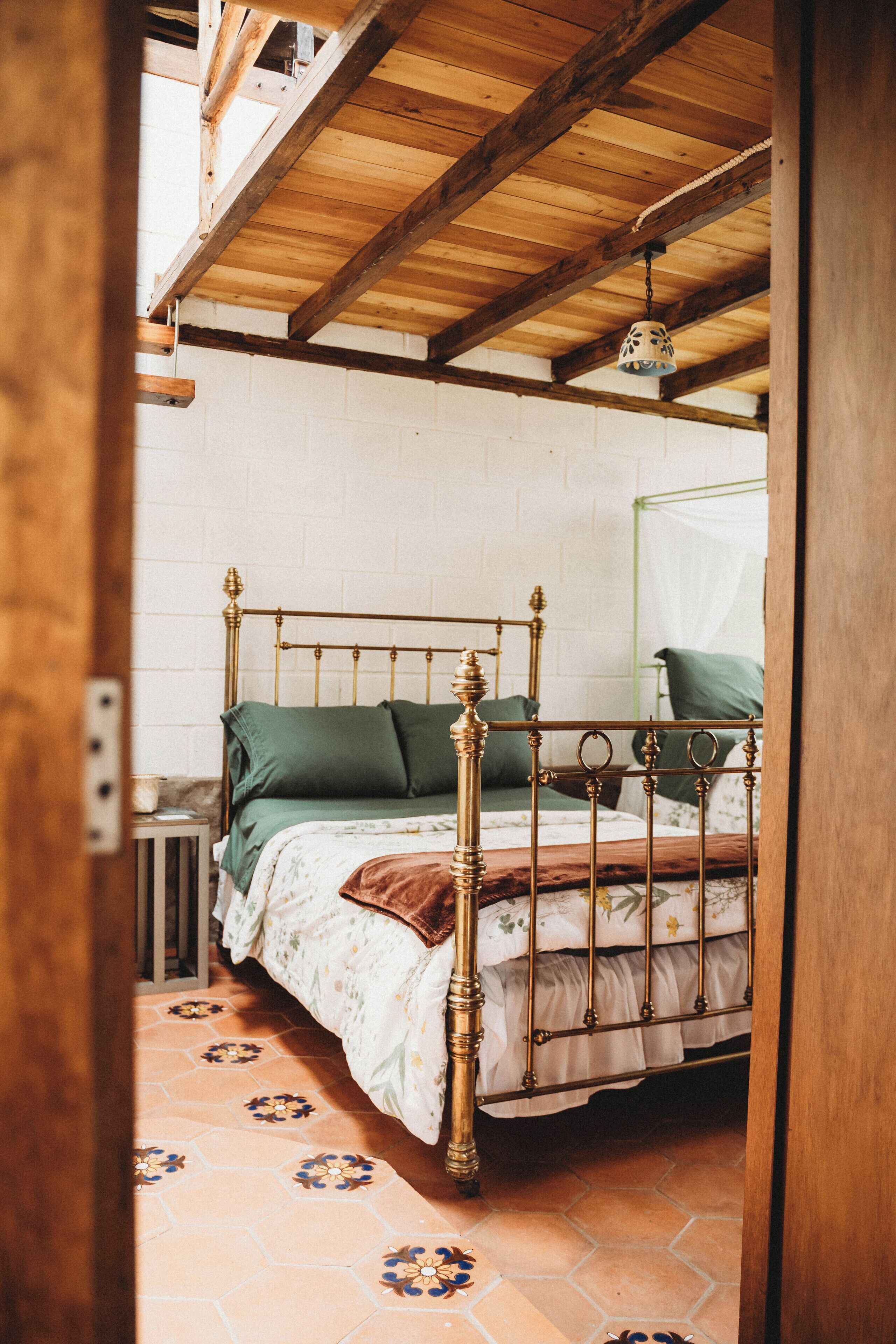 A room with a wooden ceiling, a bed with a metal frame, a green pillow, and a patterned floor.