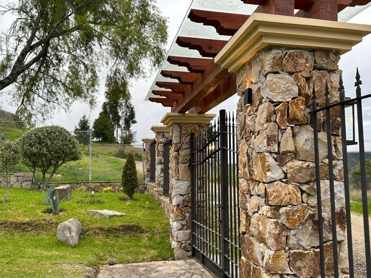 Un pilar de piedra con una reja de metal, una estructura de pérgola y un área verde con árboles y arbustos.