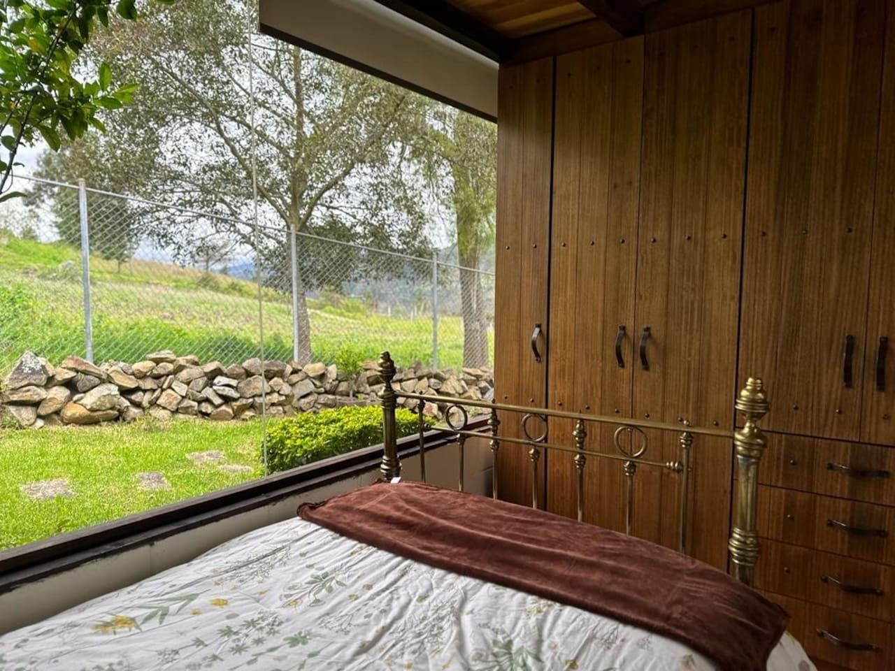 A bedroom with a large window, a bed with a brown blanket, and wooden cabinets.