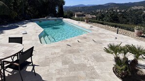 Una piscina rectangular con agua cristalina, rodeada de baldosas de piedra. En primer plano, hay una mesa y una silla, y al fondo se ve un pueblo y colinas.