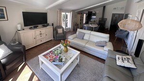Un salón amplio con un sofá blanco, una mesa de centro con revistas, un televisor y un comedor al fondo.
