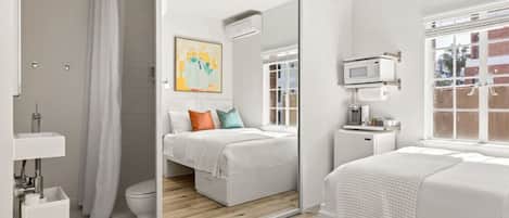 Un dormitorio moderno con un espejo grande, una mesita de noche con lámpara, una ventana con vistas y una pequeña kitchenette.