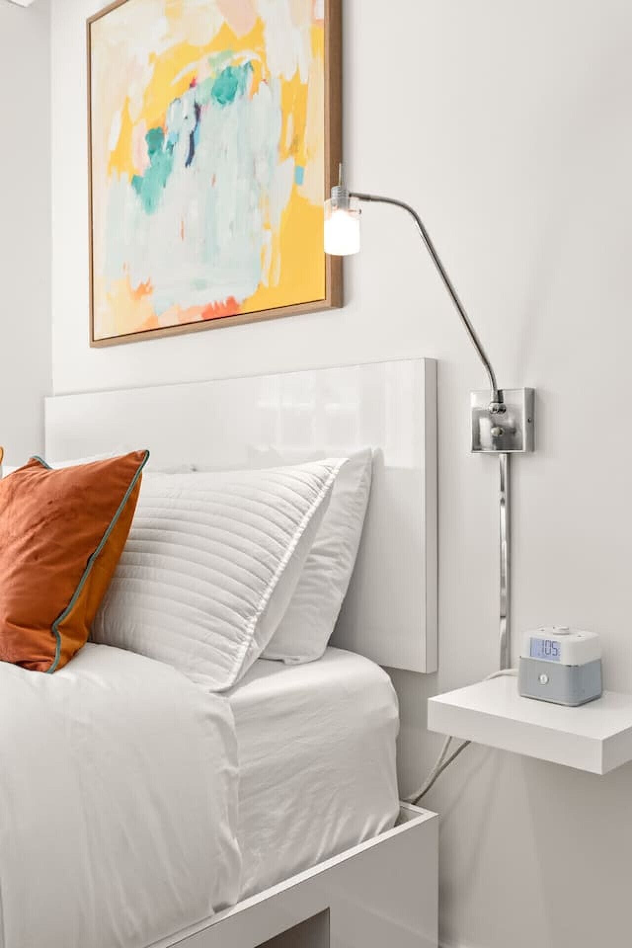 Een slaapkamer met een bed, een nachtkastje met een lamp, een digitale klok en een abstract schilderij aan de muur.
