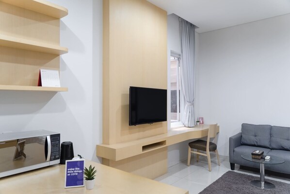 A modern living room with a flat-screen TV mounted on a wooden wall, a grey sofa, a small round coffee table, and a desk with a chair. - Cove MK House Senopati (Jakarta)