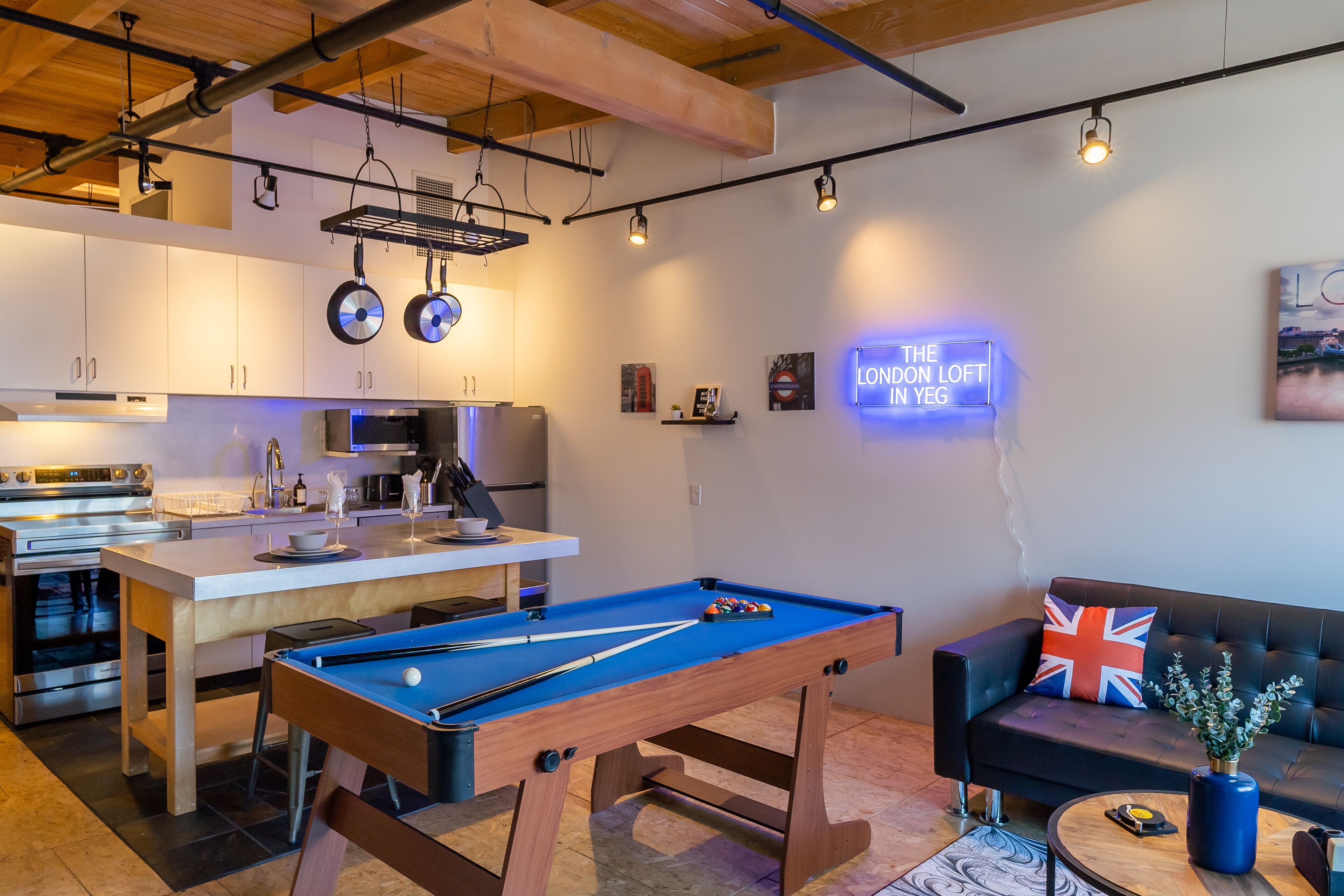 Une cuisine moderne avec des meubles blancs, un îlot central et une table de billard dans un espace de vie comprenant un canapé et un néon.