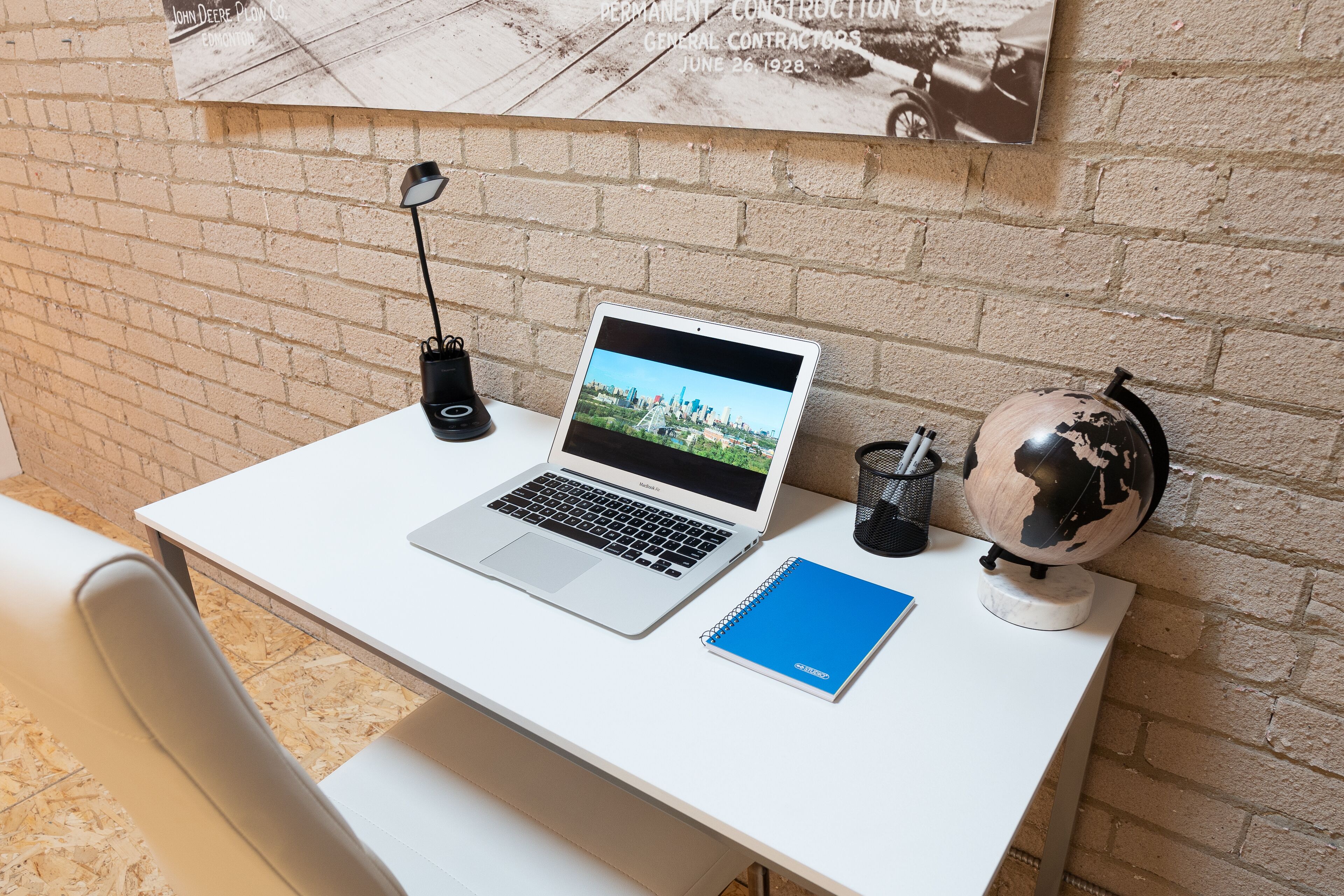 A white desk with a laptop, a blue notebook, a desk lamp, and a globe.