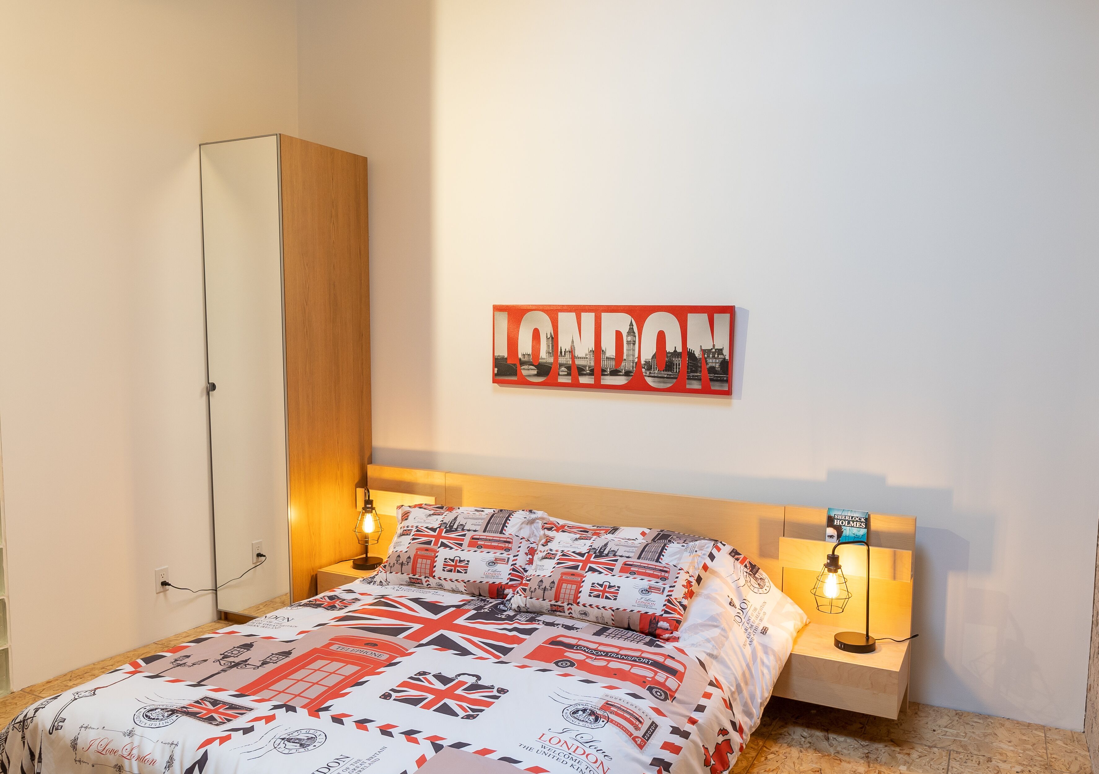 Un dormitorio con una cama que tiene un edredón con motivos de Londres, un armario de madera, un letrero en la pared que dice 'LONDON' y lámparas de noche.