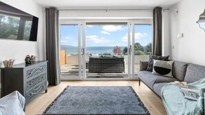 A modern living room with a grey sofa, a black rug, and a view of the sea through large glass doors. - Osprey 3, The Cove - Sea view - On site pool (Brixham)