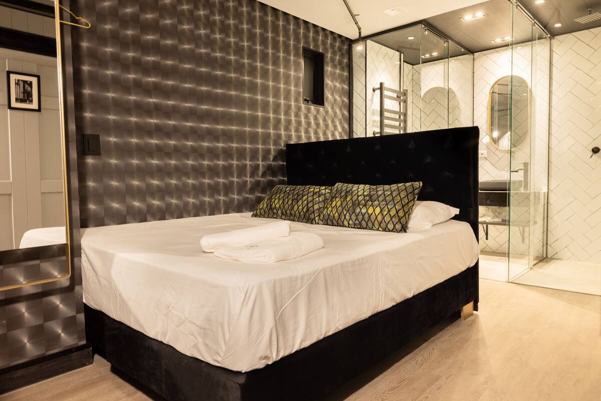 A modern bedroom with a large bed, a dark headboard, and a glass shower enclosure.