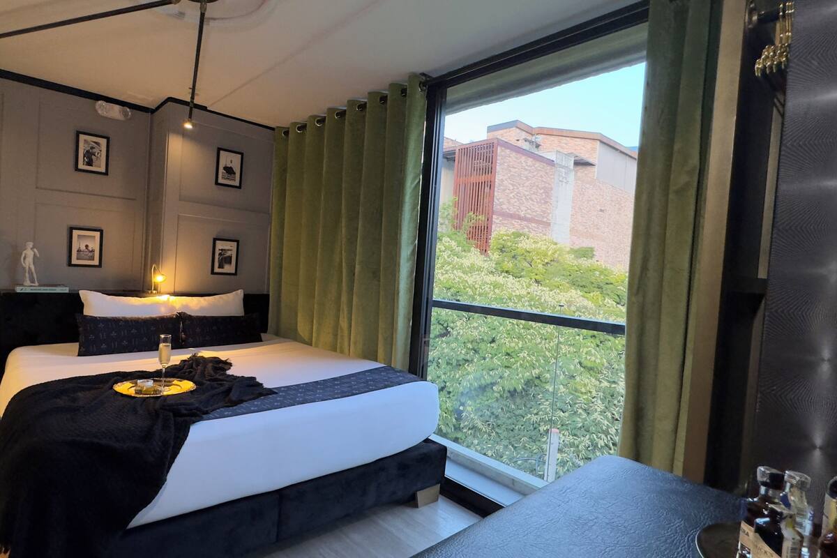 A modern hotel room with a large bed, a bedside table, a hanging robe, and a view of greenery and buildings outside.