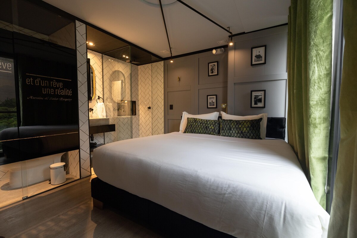 A modern hotel room with a large bed, a glass partition, a bathroom with a mirror, and framed pictures on the wall.