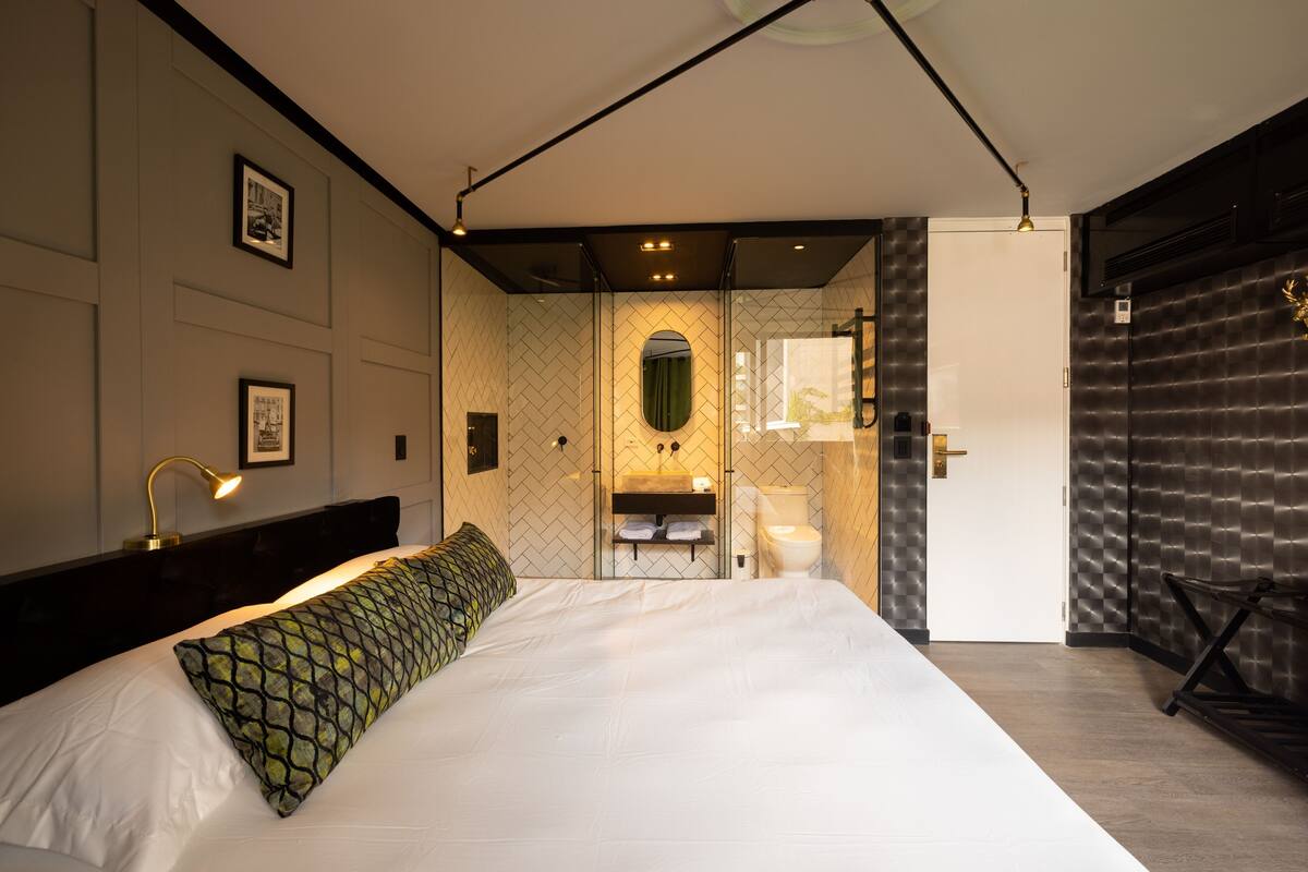 A modern hotel room with a large bed, a bathroom with a mirror, and a door leading to another room.