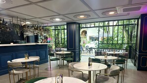 En restaurantindretning med runde borde og stole, et barområde med vinflasker, og store vinduer med udsigt til gaden.