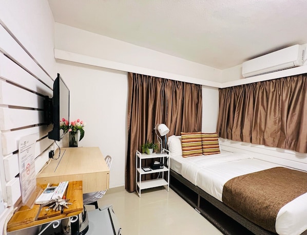 A hotel room with a bed, a desk with a computer, a chair, a TV mounted on the wall, and an air conditioning unit.