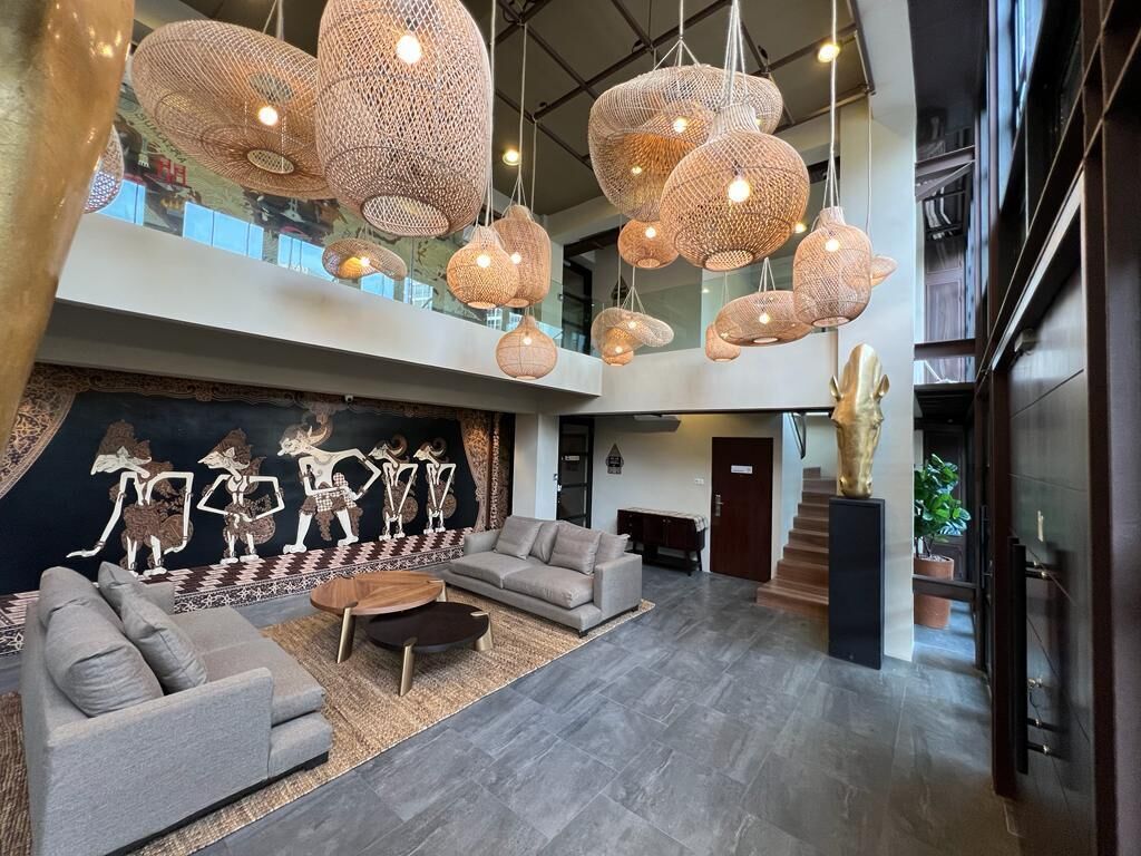 A spacious lobby with a high ceiling, multiple hanging wicker light fixtures, a large abstract mural, a grey sofa, and a staircase leading to an upper level.