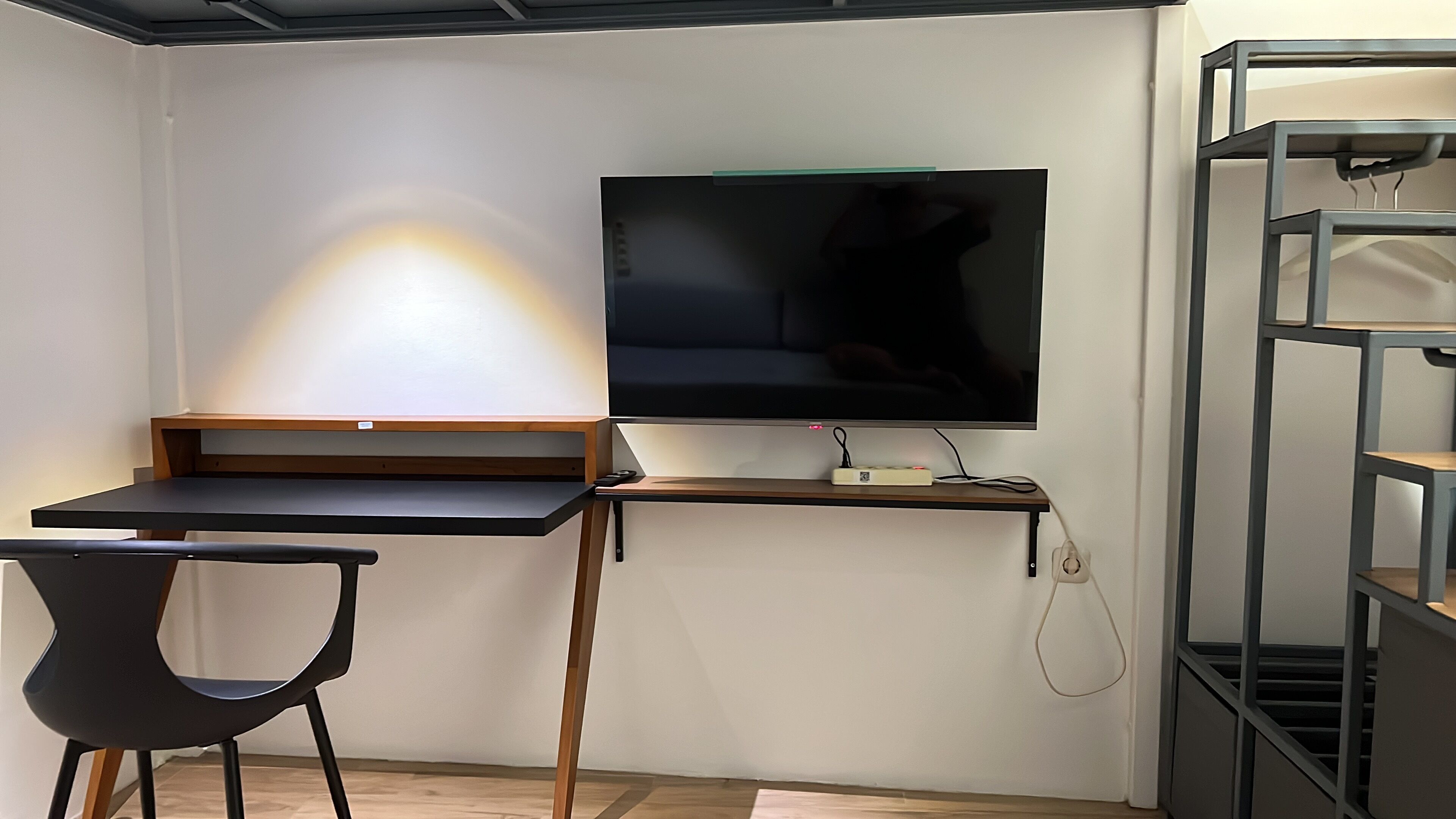Een compacte kamer met een bureau, een stoel, een flatscreen televisie aan de muur en een wandplank aan de rechterkant.
