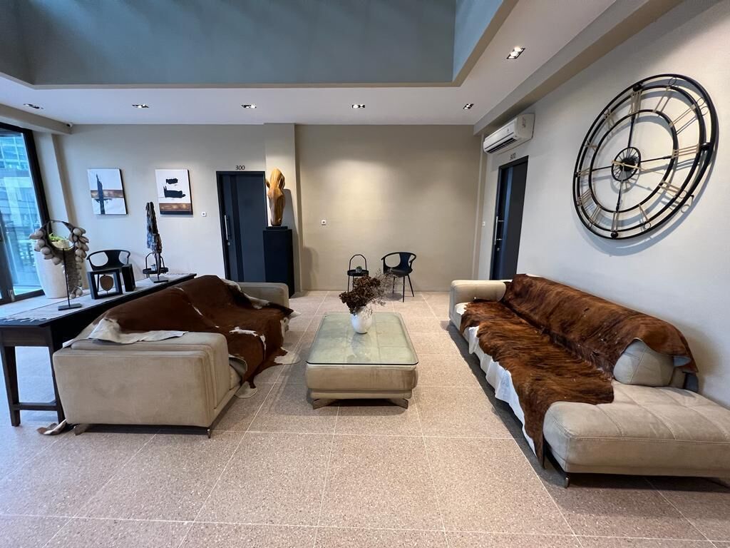 Een moderne woonkamer met een grote bruine bank, een kleinere bank en een glazen salontafel. Achtergrond toont een grote wandklok en een deur met de tekst '300'.
