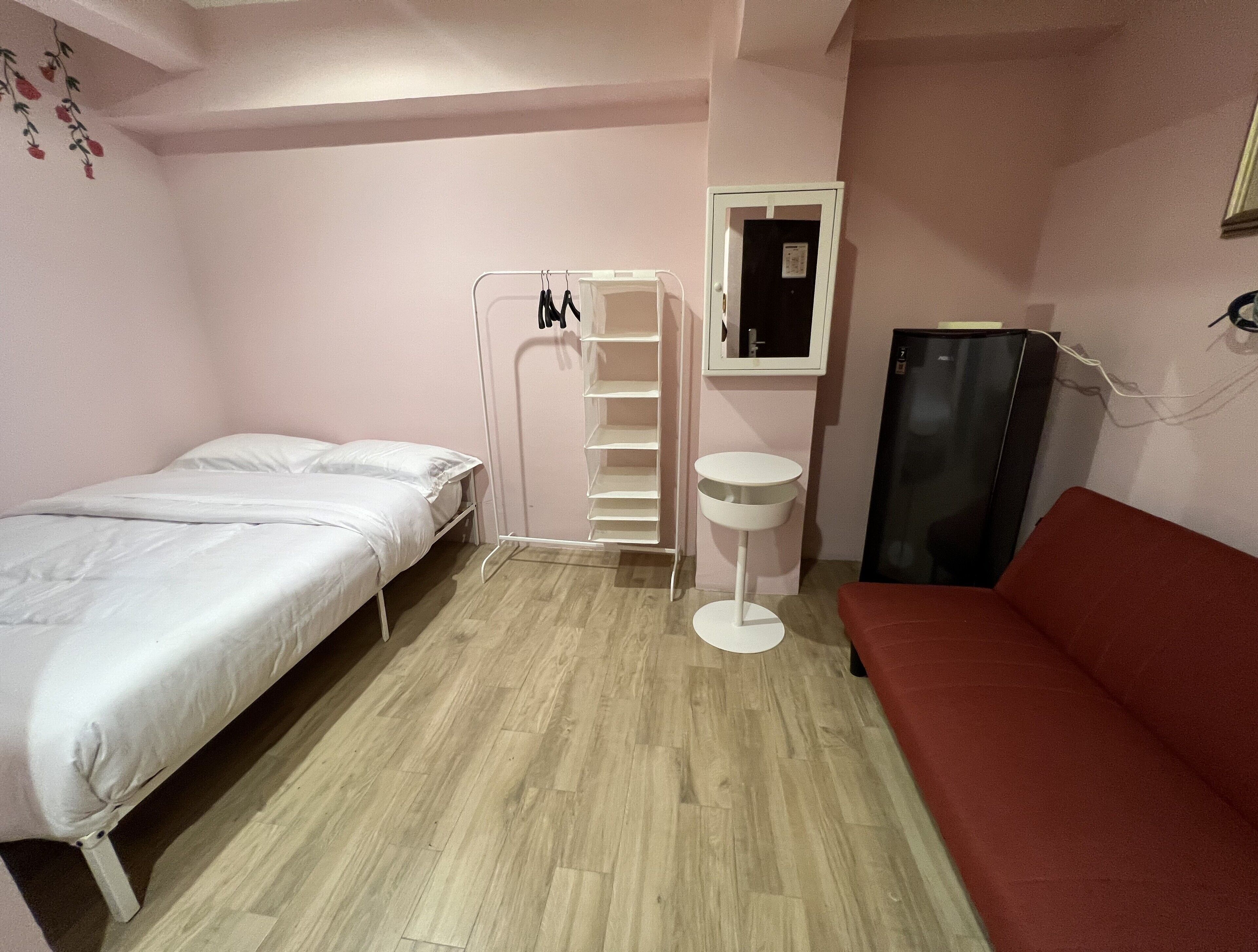 A small room with a bed, a red sofa, a white bedside table, a white wardrobe, and a small window.
