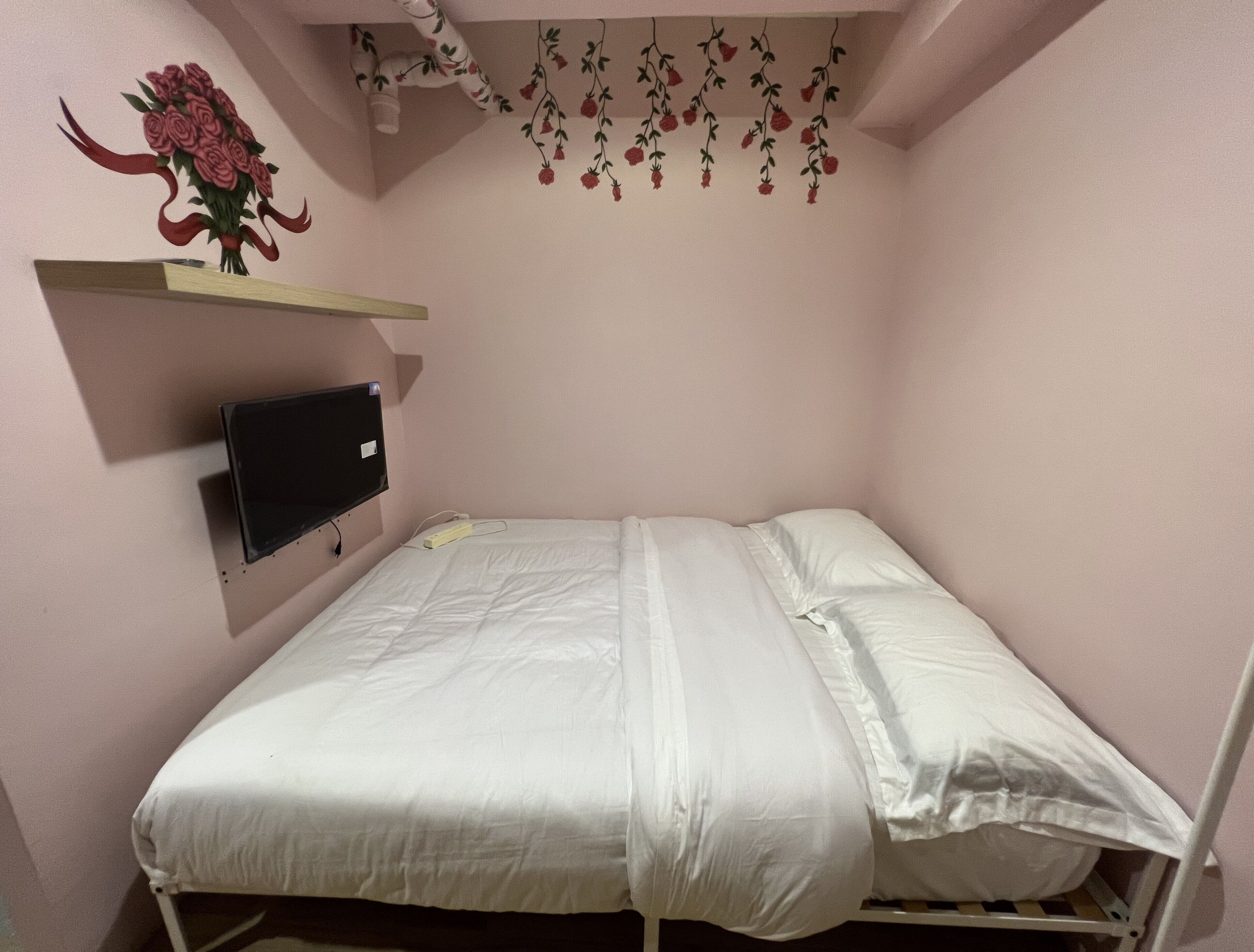 Una cama individual con ropa blanca, un televisor montado en la pared y un estante con un adorno floral.