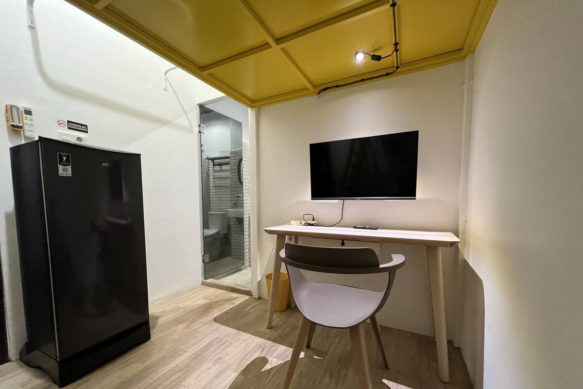 A compact room with a black refrigerator, a desk with a chair, a mounted TV, and a glass shower enclosure.