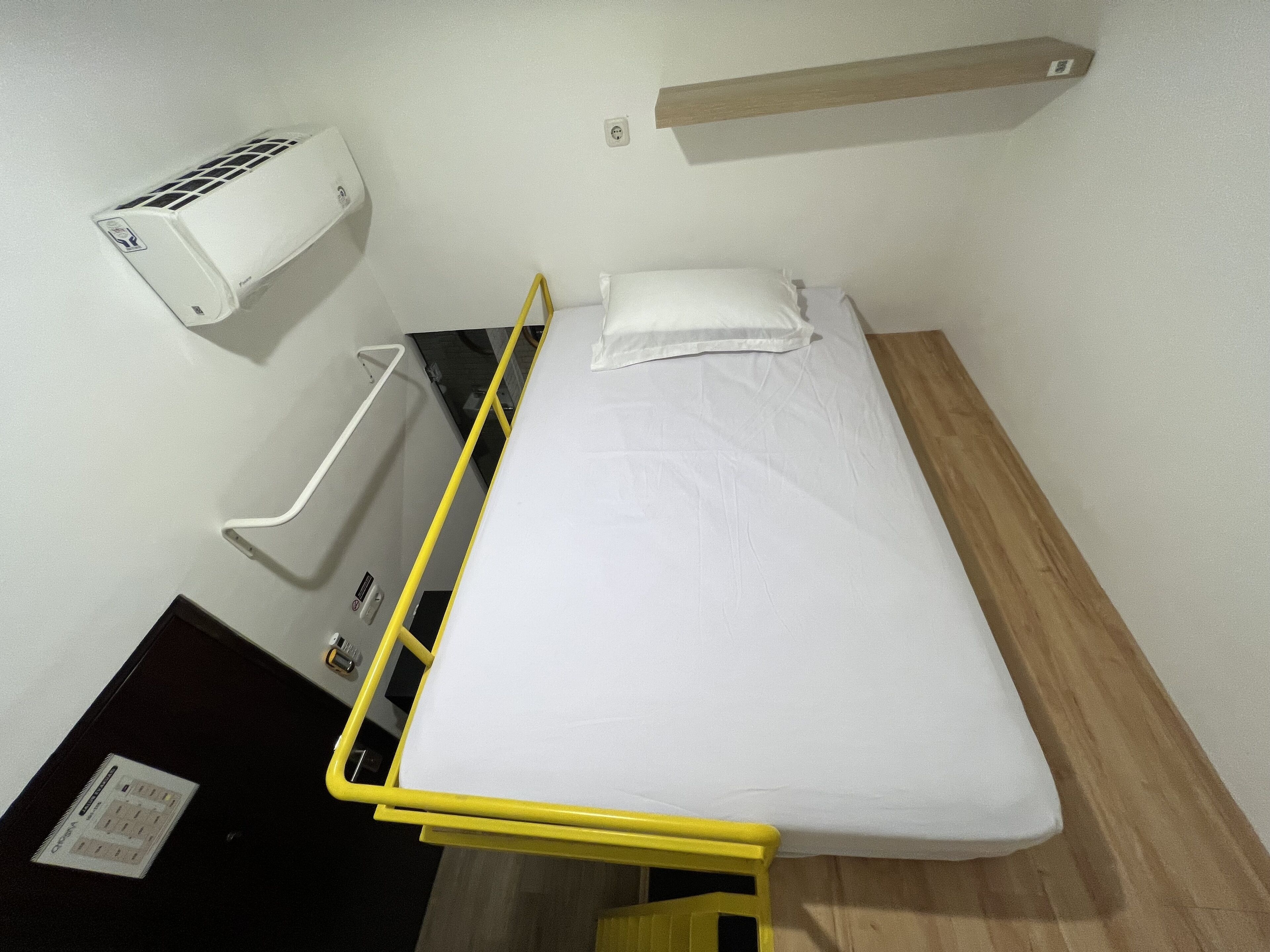 A single bed with a white mattress and pillow, a yellow metal frame, a wall-mounted air conditioner, and a wooden shelf above the bed.