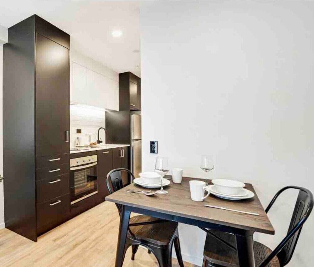 Een moderne keuken met donkere kastjes, een eettafel gedekt met borden, glazen en bestek, en een kleine koelkast.