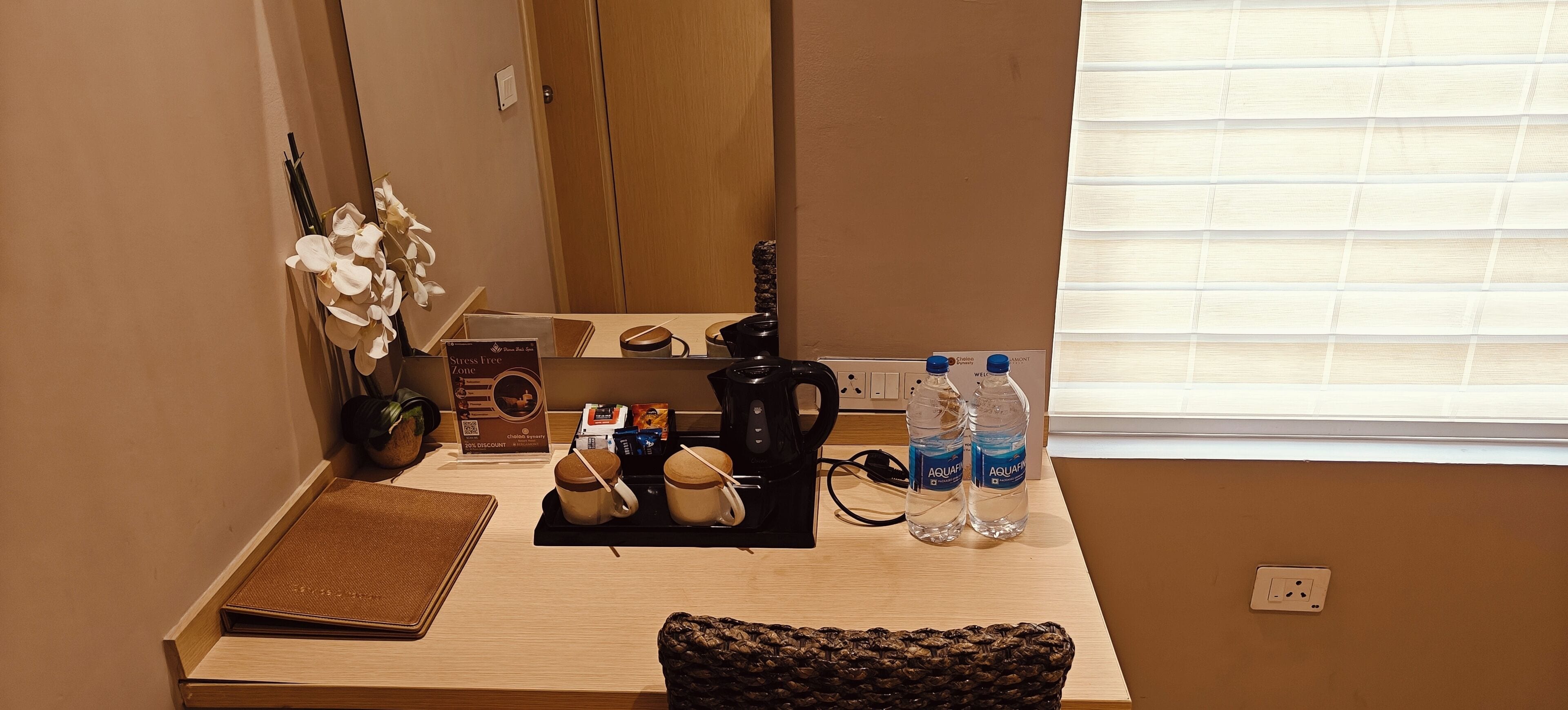 Une chambre d’hôtel avec un bureau en bois, une bouilloire noire, deux bouteilles d’eau, une petite plante et un panneau indiquant une zone propice à la détente.
