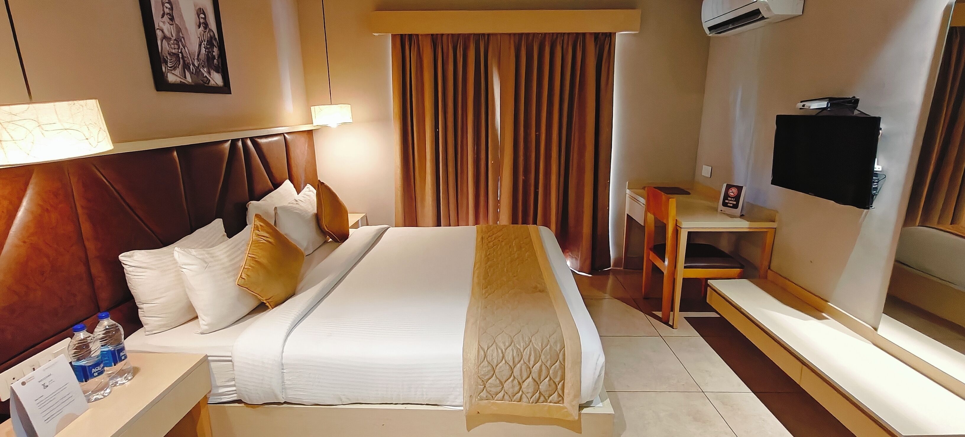 A hotel room with a bed, bedside table, lamp, curtains, and a TV.