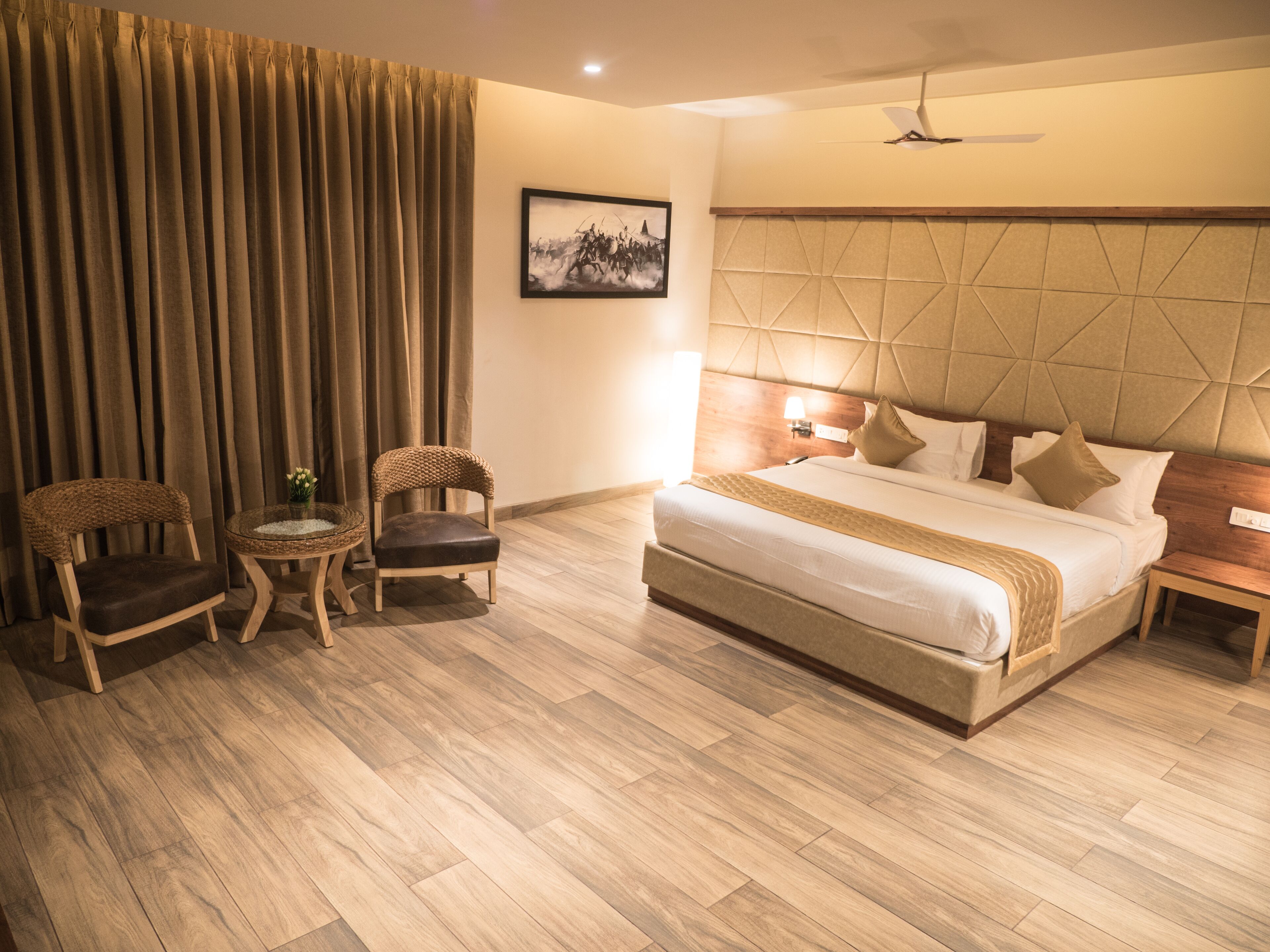 Habitación de hotel con una cama grande, dos sillas de mimbre, una mesita con un jarrón y un cuadro en la pared.