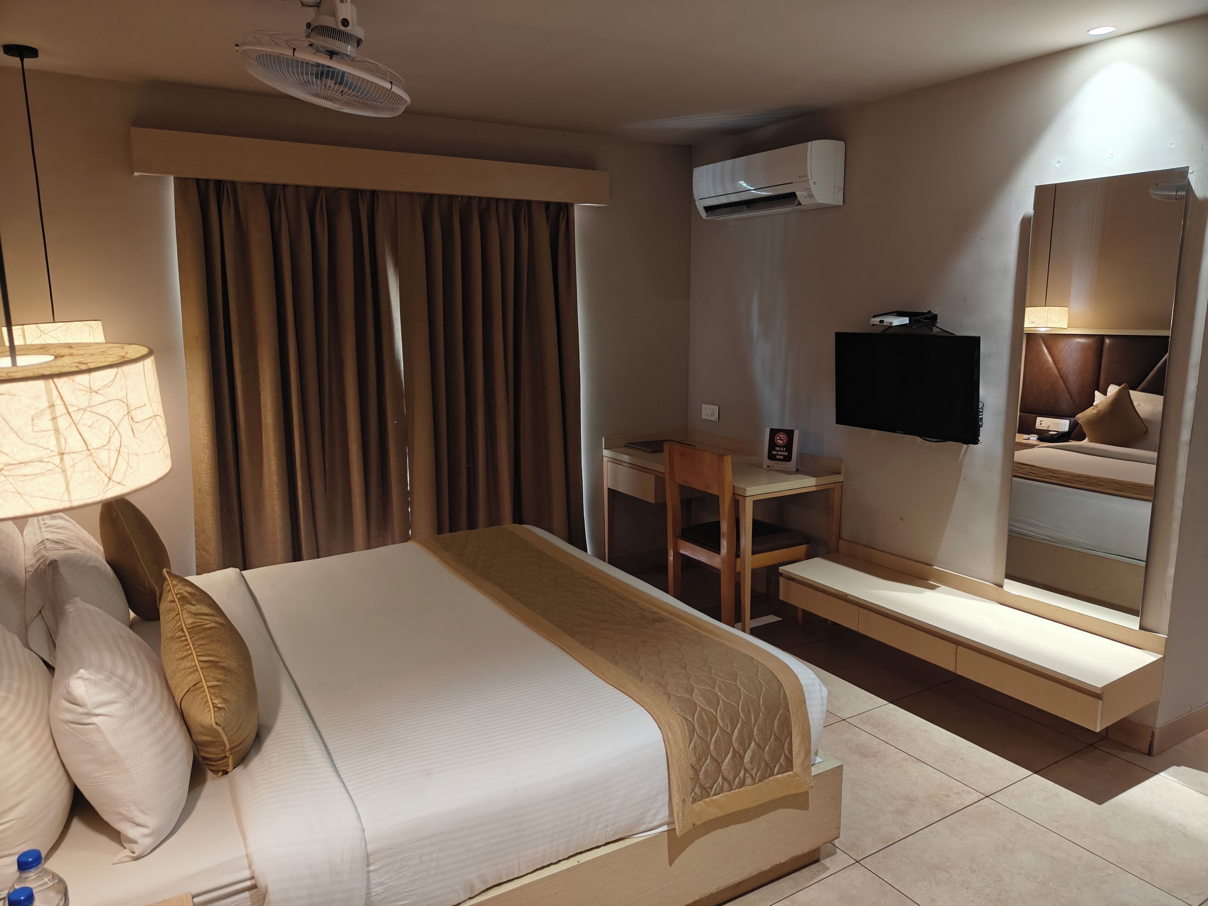 Habitación de hotel con cama, escritorio, silla, aire acondicionado, televisor y espejo.