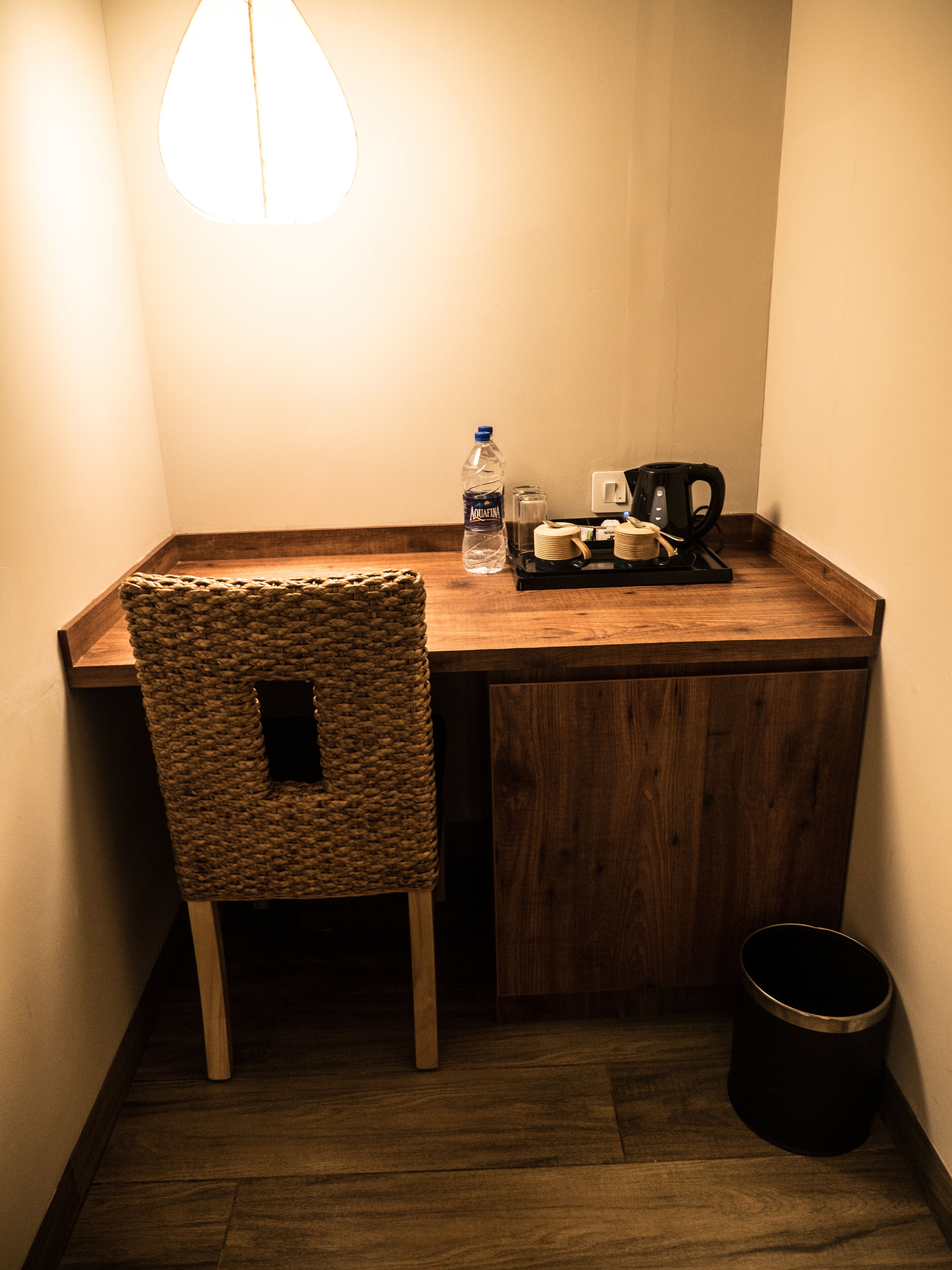 Un rincón pequeño y acogedor con un escritorio de madera, una silla tejida, una botella de agua, una cafetera y un cubo de basura.