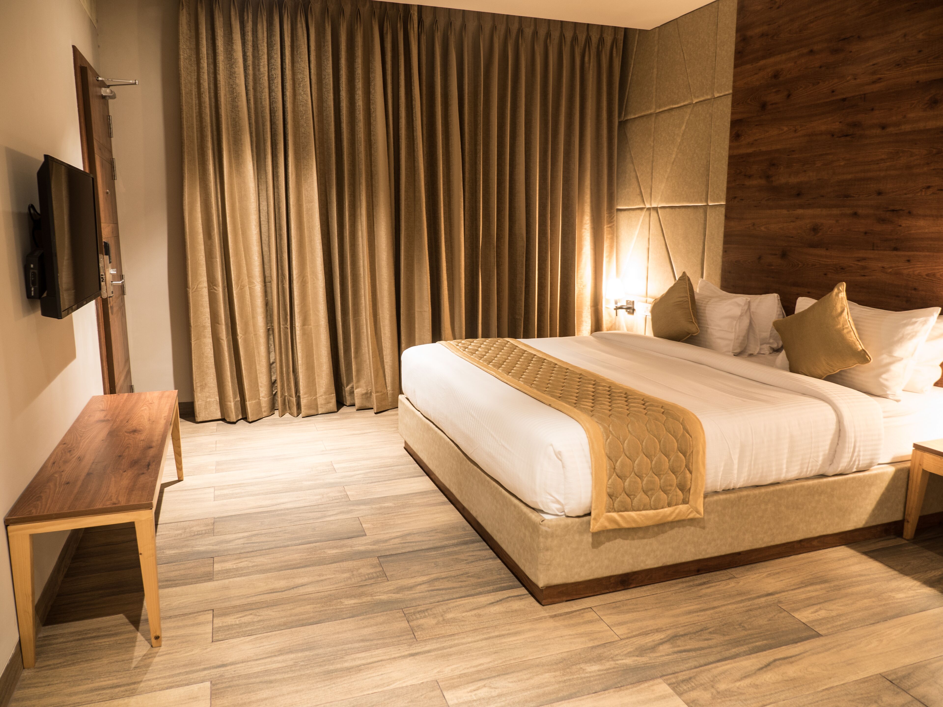 A modern hotel room with a large bed, wooden floor, a bench, and a TV on the wall.