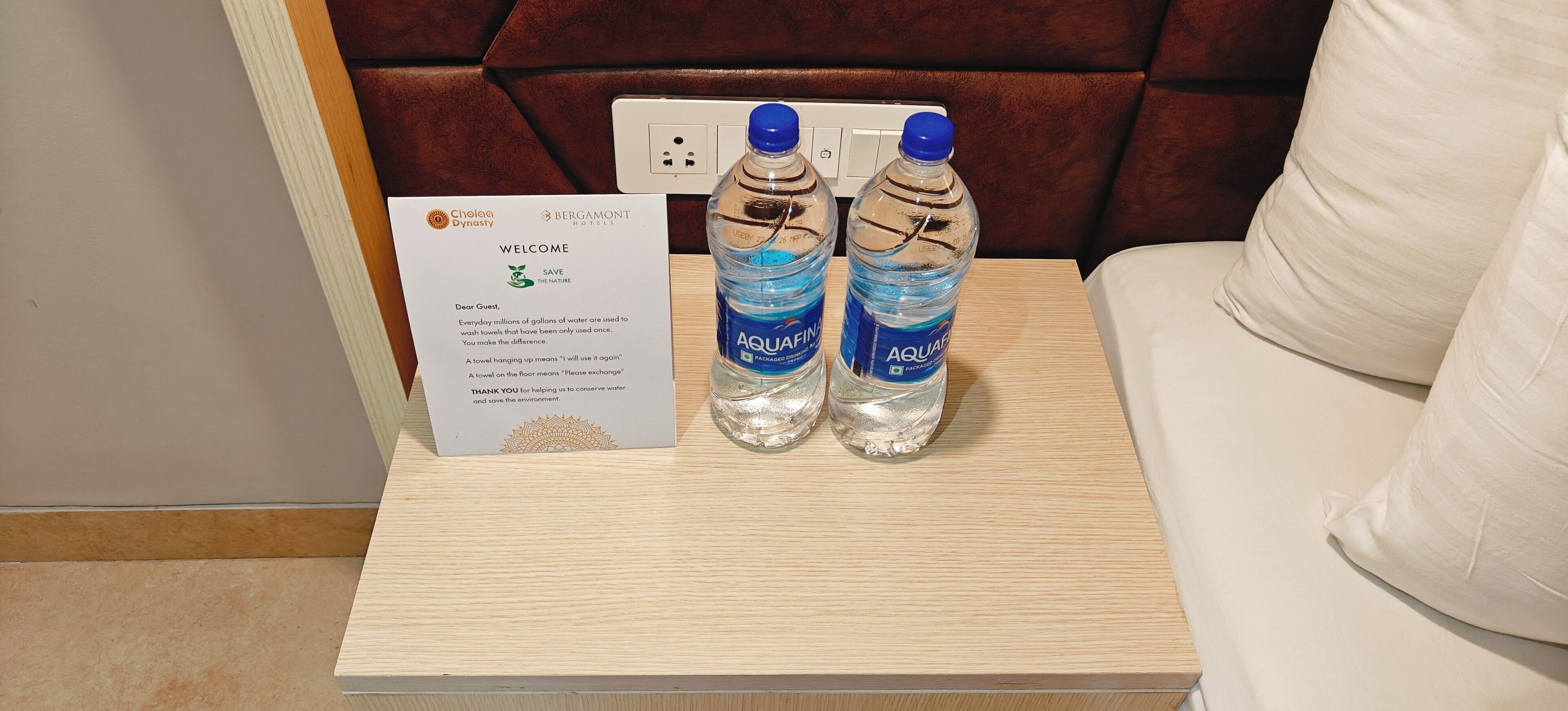 Une pancarte d’accueil affichant un message sur la nécessité d’économiser l’eau, ainsi que deux bouteilles d’eau Aquafina, sont posées sur une table de chevet en bois.