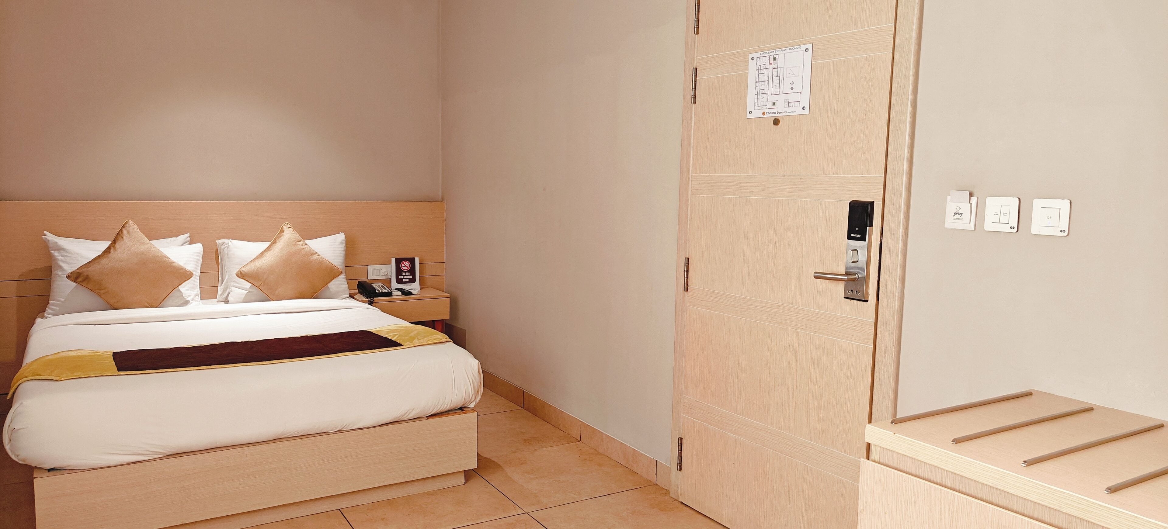A hotel room with a bed, a nightstand, a wall-mounted emergency exit plan sign, a door with a handle, and a small shelf with items.