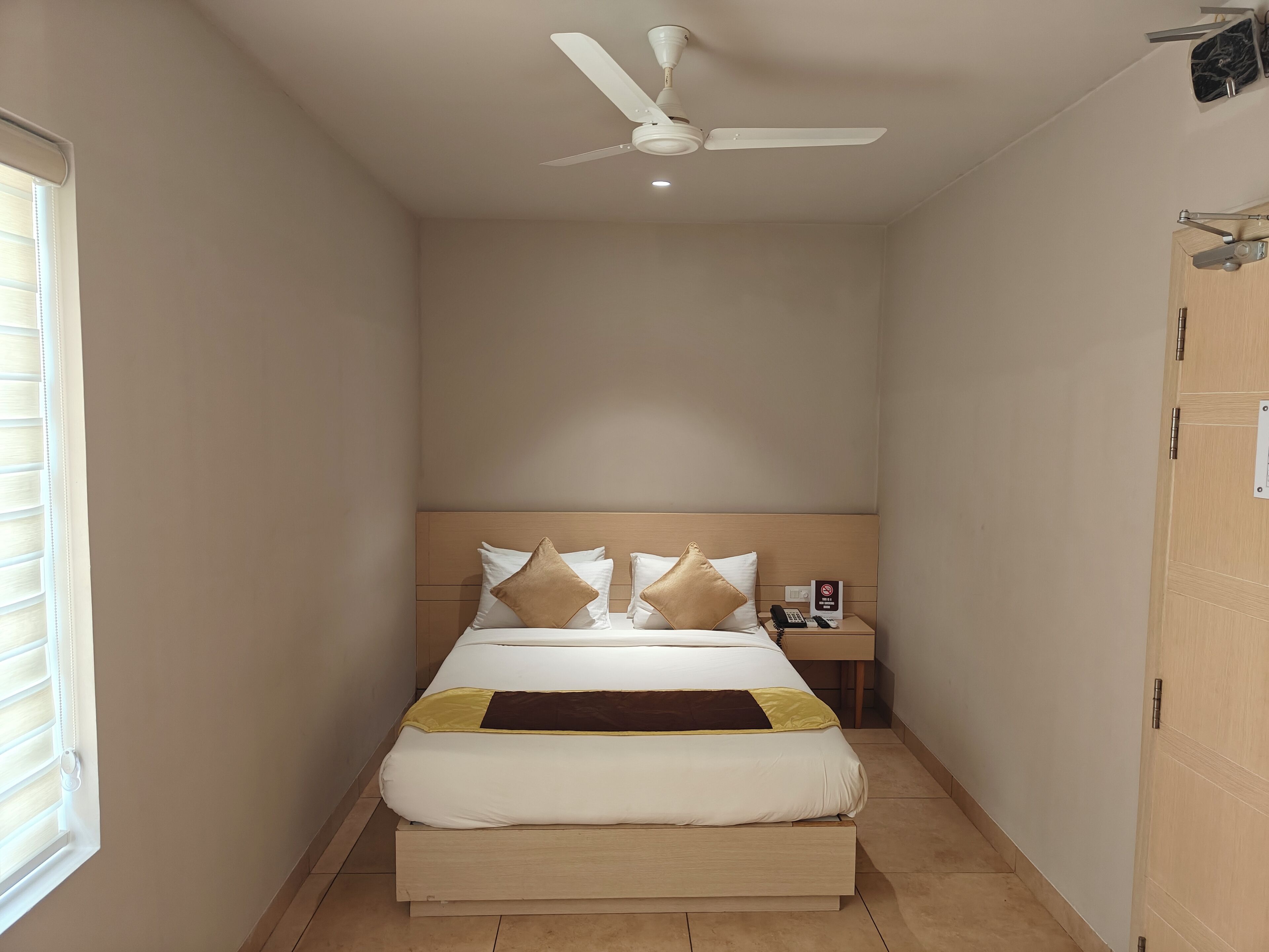 Una cama individual con cabecera de madera, una mesita de noche con un teléfono y un cartel, y un ventilador de techo.