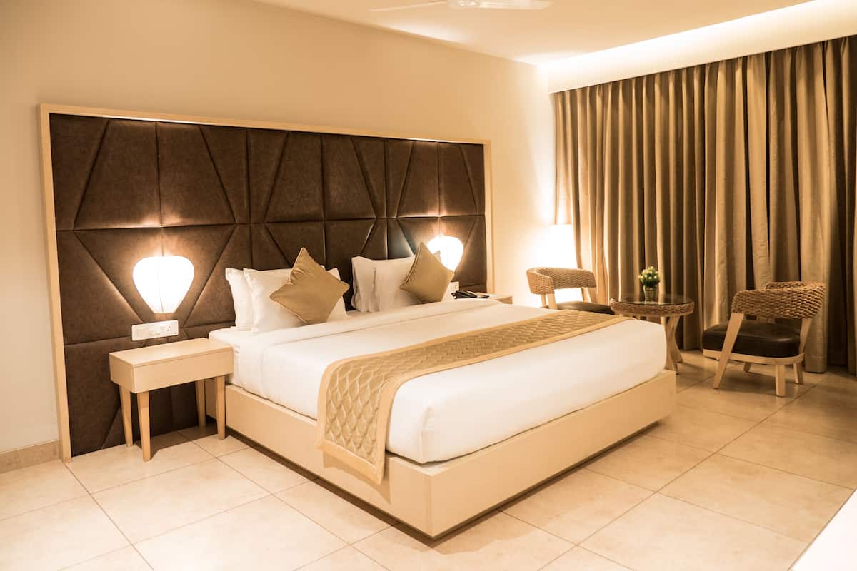 Une chambre d’hôtel moderne avec un grand lit, une tête de lit ornée d’un motif géométrique, une table de chevet avec une lampe, une petite table d’appoint avec une plante et un fauteuil.