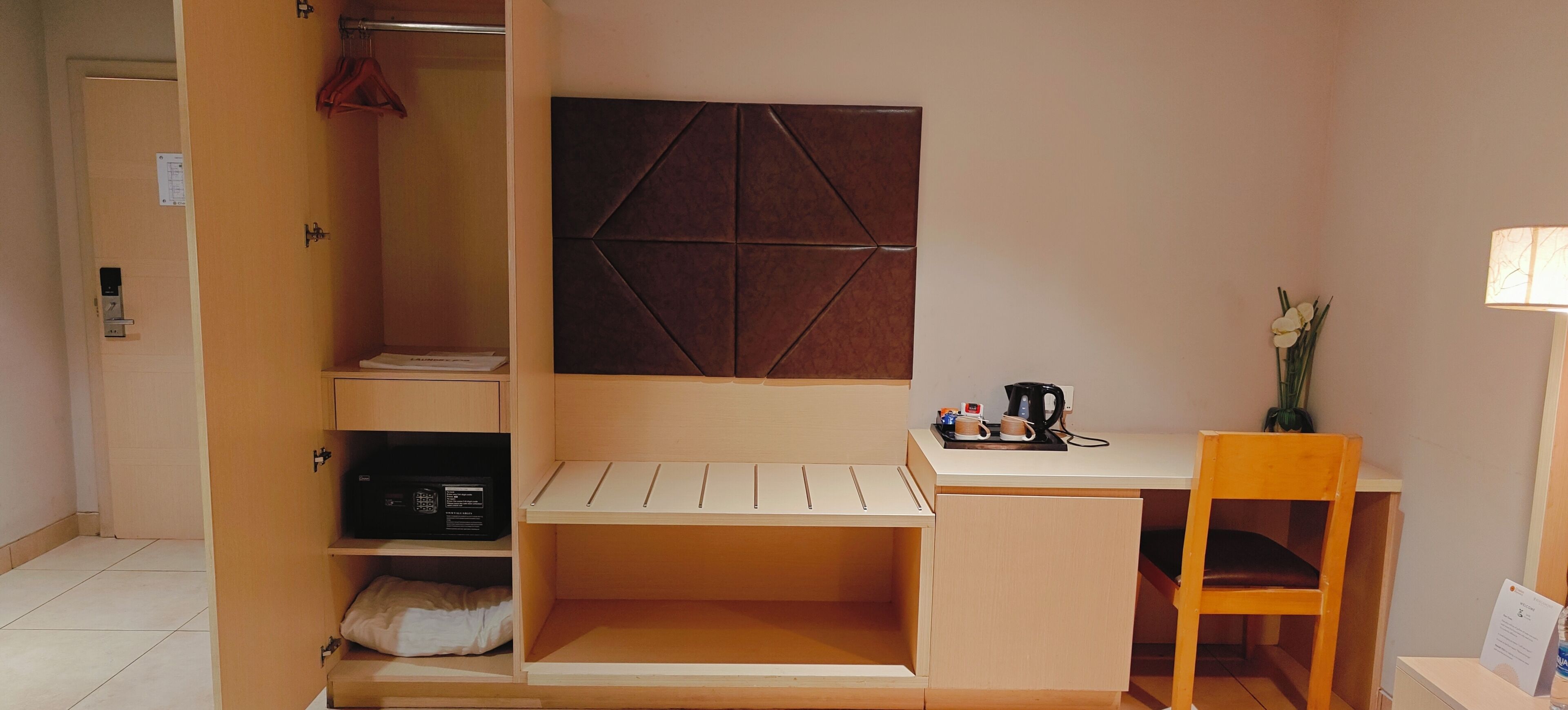 A compact room with a bed, a desk, and a chair. There is a wall-mounted panel and a small table with a kettle and a cup.