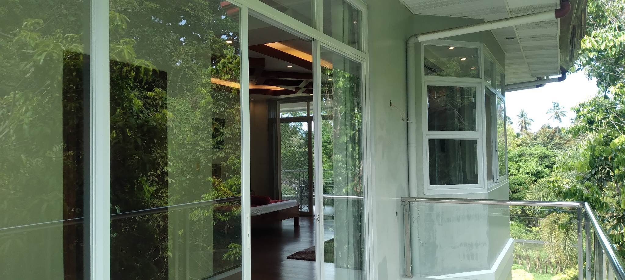 A modern building with large glass windows reflecting greenery, a balcony with a metal railing, and a visible interior with a bed and a table.