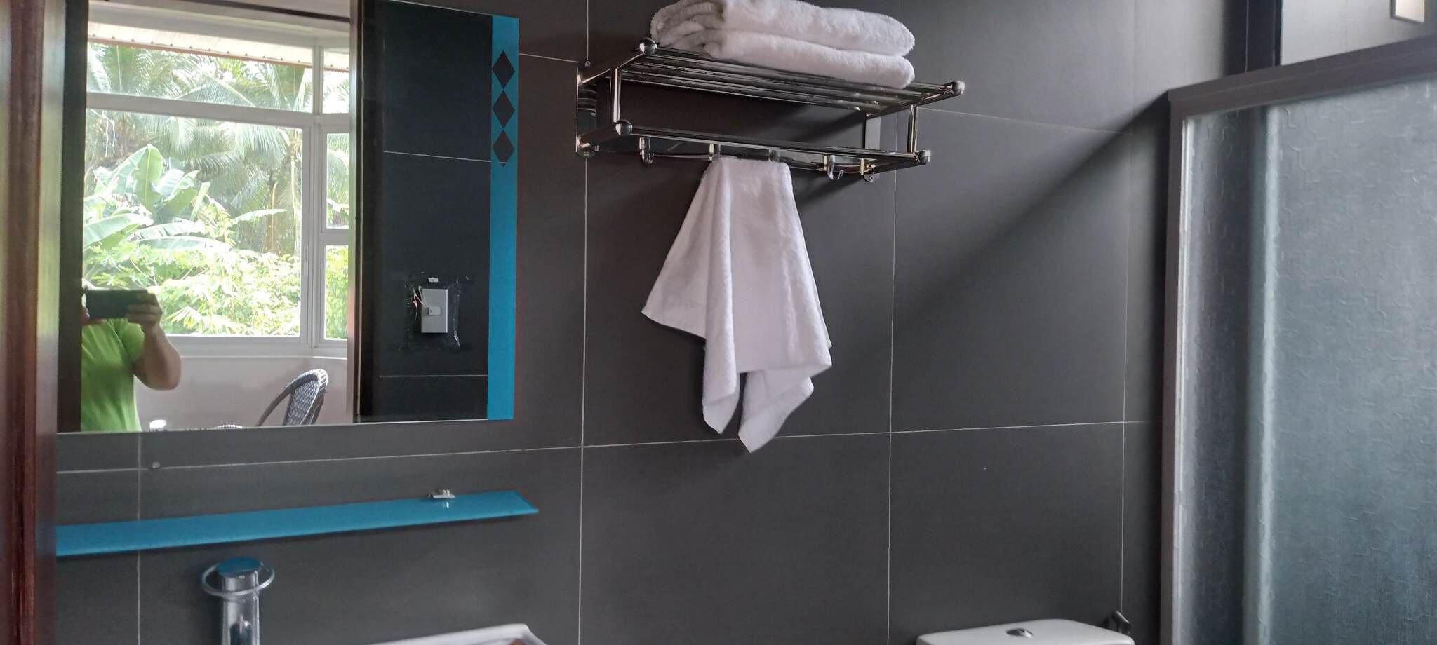 Egy modern fürőszoba, melyben egy nagy tükör, egy törölközőtartó, rajta szépen összehajtogatott törölközők, egy kék törölközőrúd és egy ablak található, melyen át zöld növényzet látható.