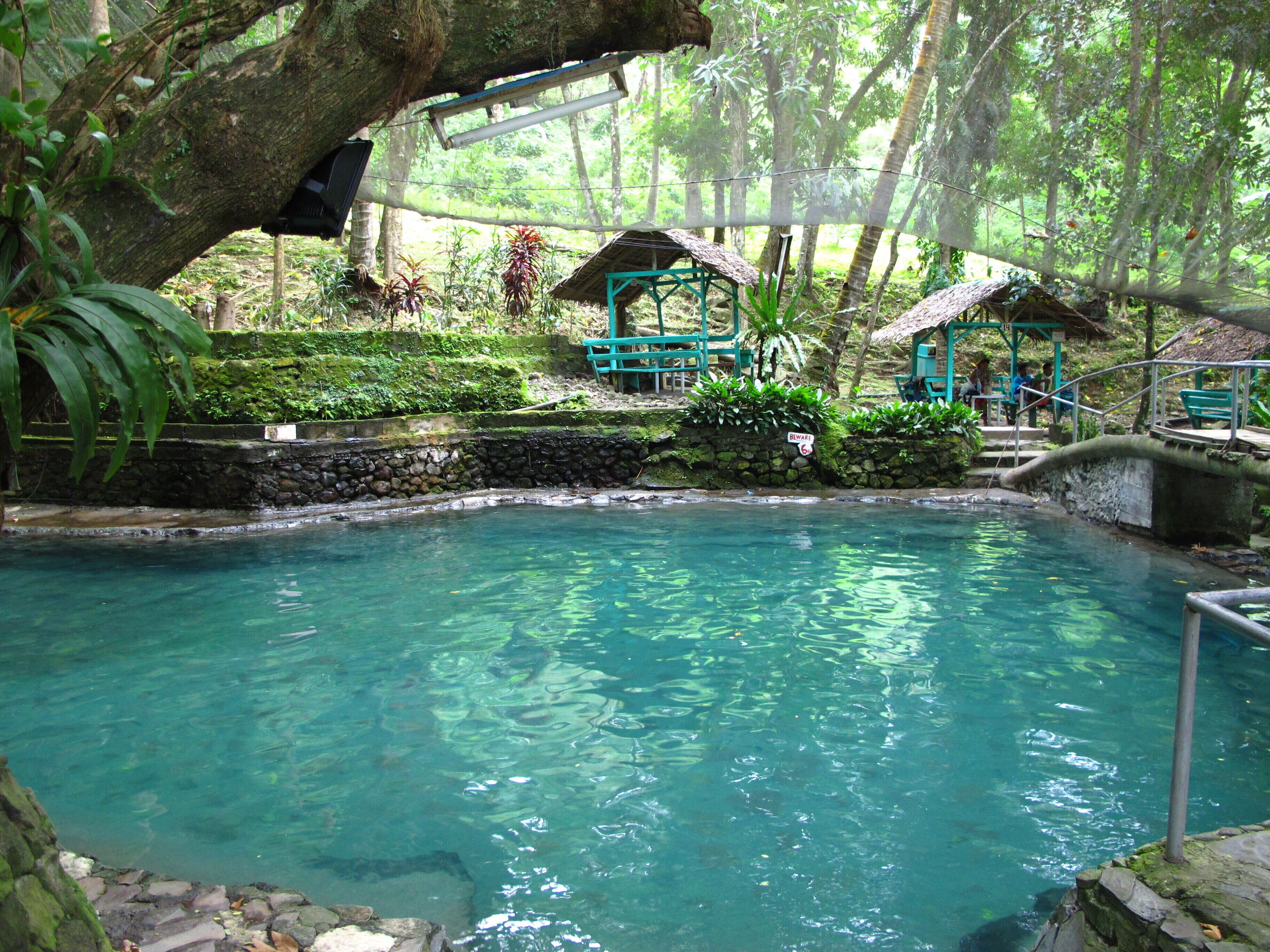 Una piscina cristalina rodeada de muros de piedra y exuberante vegetación tropical. Se observan construcciones con techo de paja y una zona de asientos de piedra.